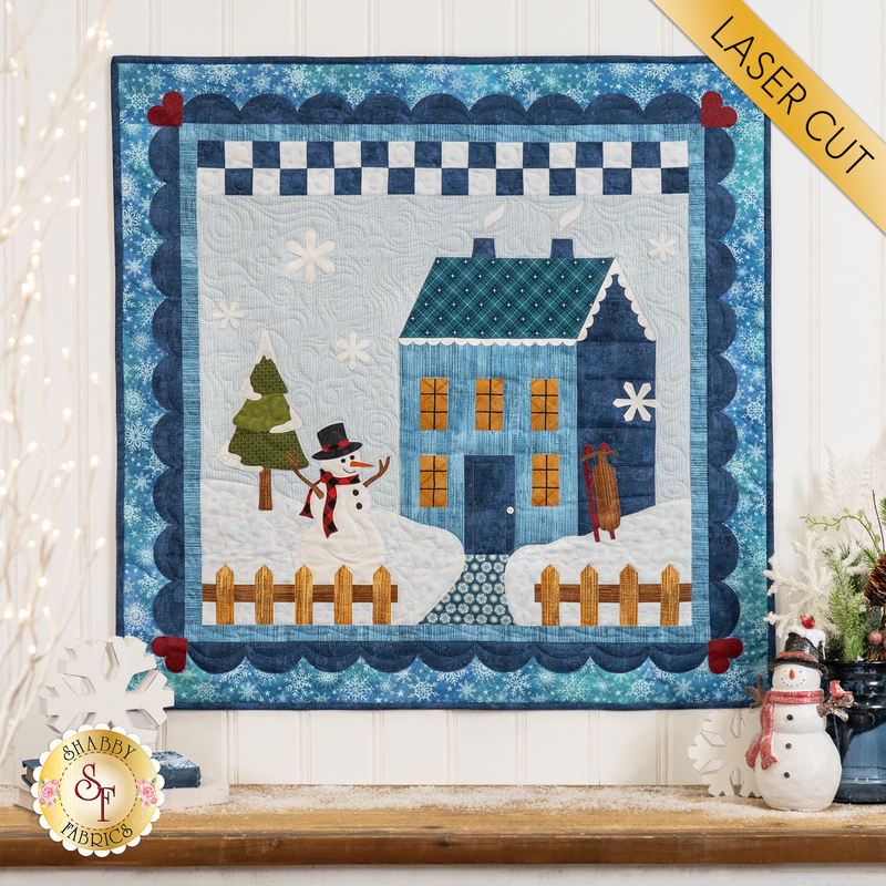 A quilted wall hanging featuring a blue house with a snowman and a tree in a winter scene. The background has a border with a checkered pattern and snowflakes. There is a small wooden fence in front of the house. A banner in the top corner reads LASER CUT.