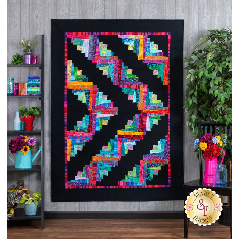 A vibrant quilt featuring a geometric pattern in bright colors, predominantly arranged in a log cabin style, set against a black background. The quilt is displayed on a wall in a room with wooden flooring and decor items, including shelves with books and plants.