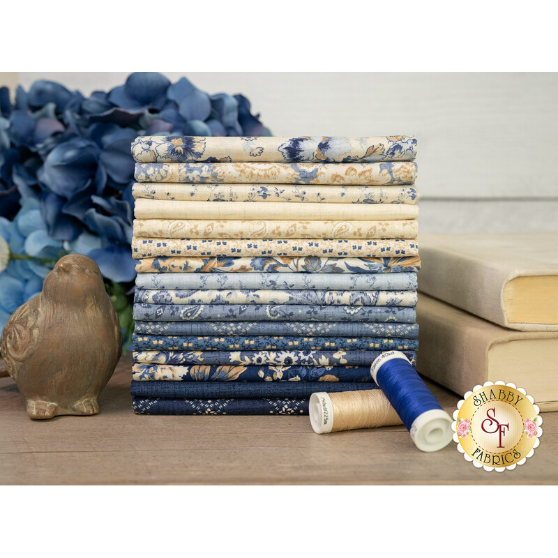 A stack of all the Radiance fabrics, in lovely shades of cream, white, and blue, with flowers, books, thread, and a bird statue.