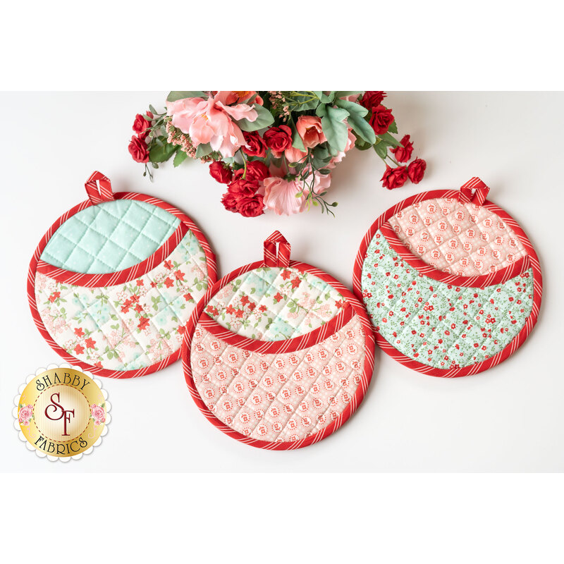 The three completed hot pads in teal, pink, and red, spread out on a white background with a bouquet of pink and red craft flowers at the top of the photo.