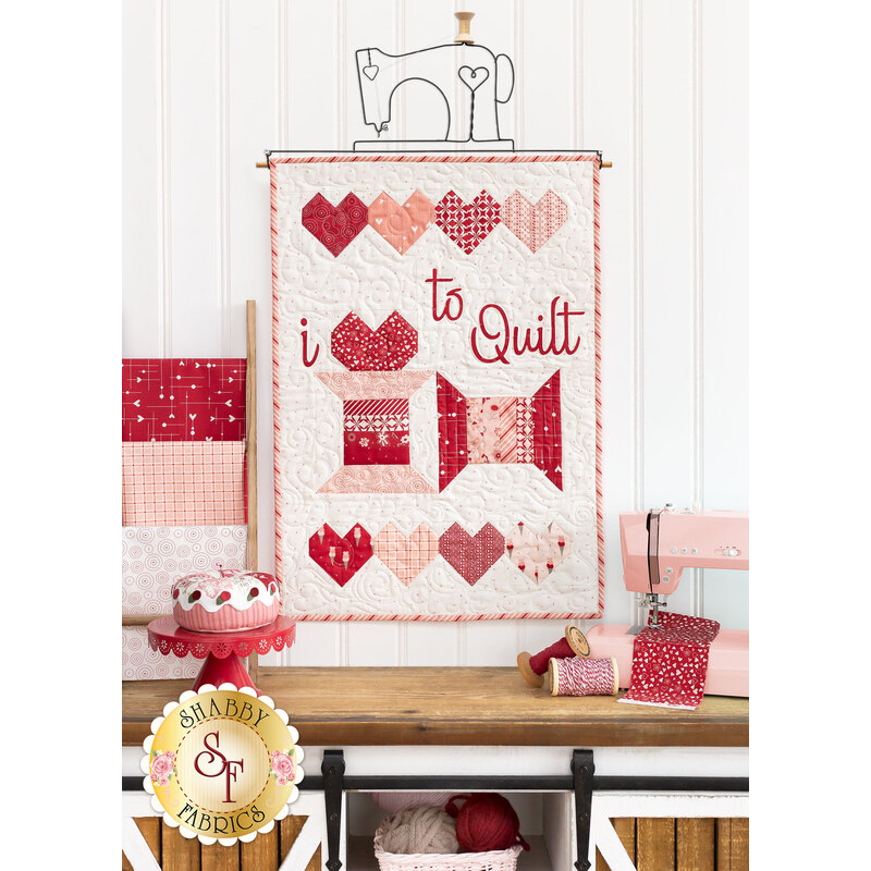 A head-on shot of the completed wallhanging, staged on a craft hanger that has decorative wirework shaped as a sewing machine. The Wallhanging is staged above a wooden work table that holds a pink sewing machine, dessert pincushion, and various threads and fabrics.