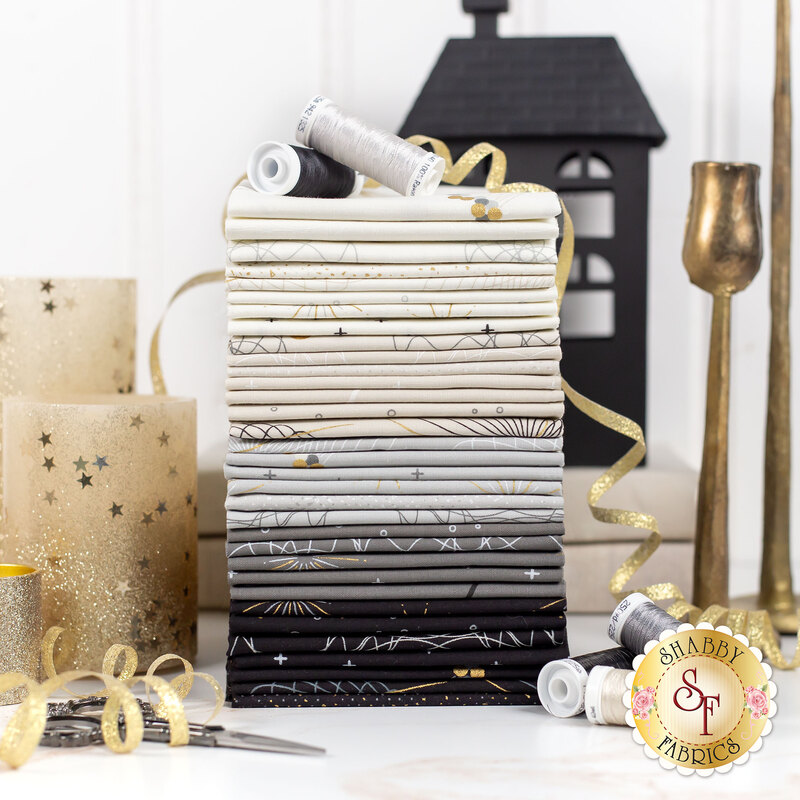  white, ecru, gray, and black fabric with gold metallic accents on a white table surrounded by gold ribbon, candles, and neutral threads