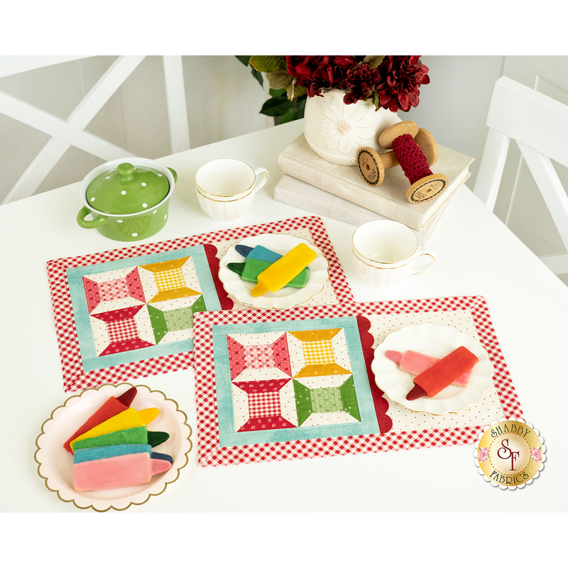 The two completed Tea & Cookies placemats for September, colored in bright yellow, green, red and pink, staged on a white table with coordinating colors and floral decor. A ceramic cup of red flowers peeks into frame from above.