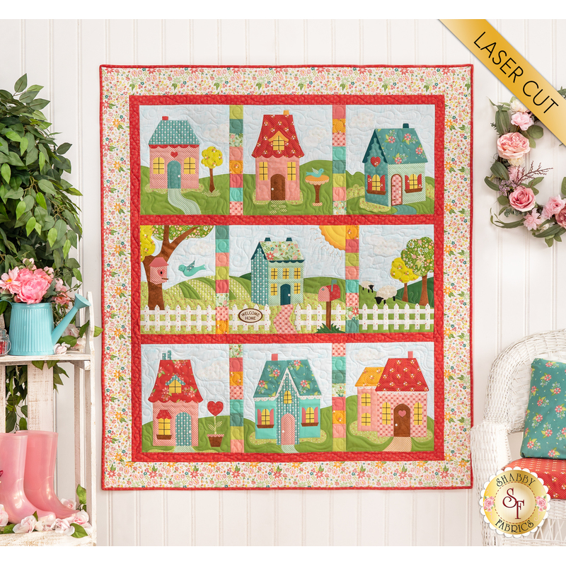 The completed Welcome Home In Spring quilt in bright, cheerful colors, hung on a paneled wall beside pink rainboots, a watering can planter with pink flowers, and a wreath of pink roses. A yellow banner in the upper right hand corner reads "Laser Cut"