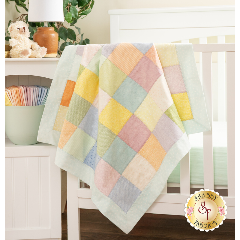 The completed patchwork receiving blanket in gentle pastel colors, artfully draped over the side of a crib, staged with coordinating white furniture, fabric, and a stuffed teddy bear.