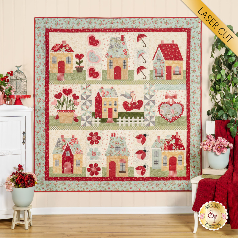 The completed English Cottages quilt, colored in warm reds, soft teal, and classic cream, with mossy green and baby pink accents. It is staged on a cream wall and bordered by coordinating furniture and housewares. A yellow banner at the top right reads "Laser Cut"