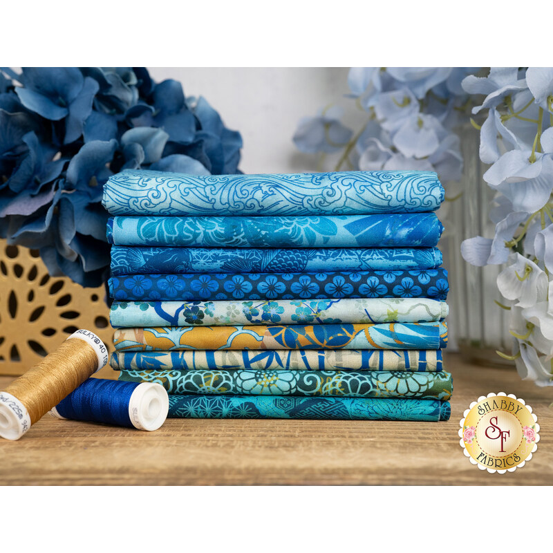 A stack of the fabrics included in the Oriental Gardens Blue FQ set, with flowers and vases and thread