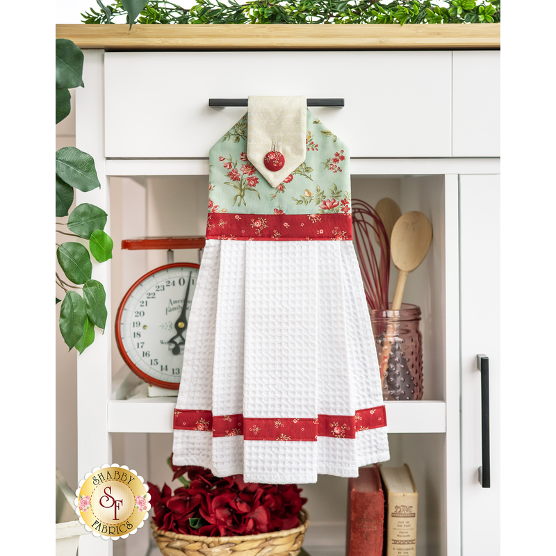 The completed hanging towel in blue, red, and cream, staged on a kitchen cart with cooking utensils, a scale, flowers, and books.