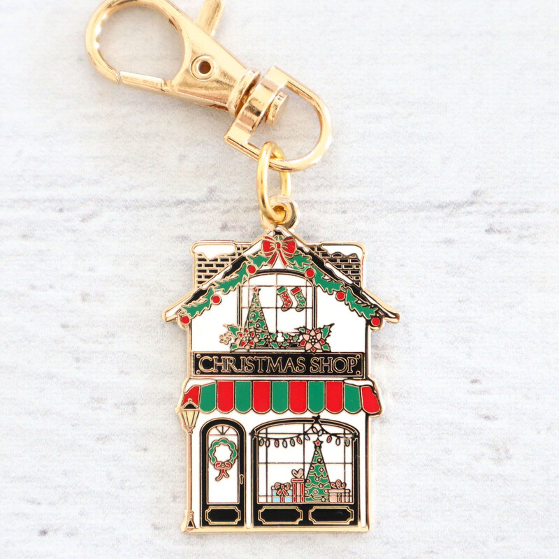 Gold keychain charm shaped like a Christmas shop, decorated with holiday ornaments and a tree.