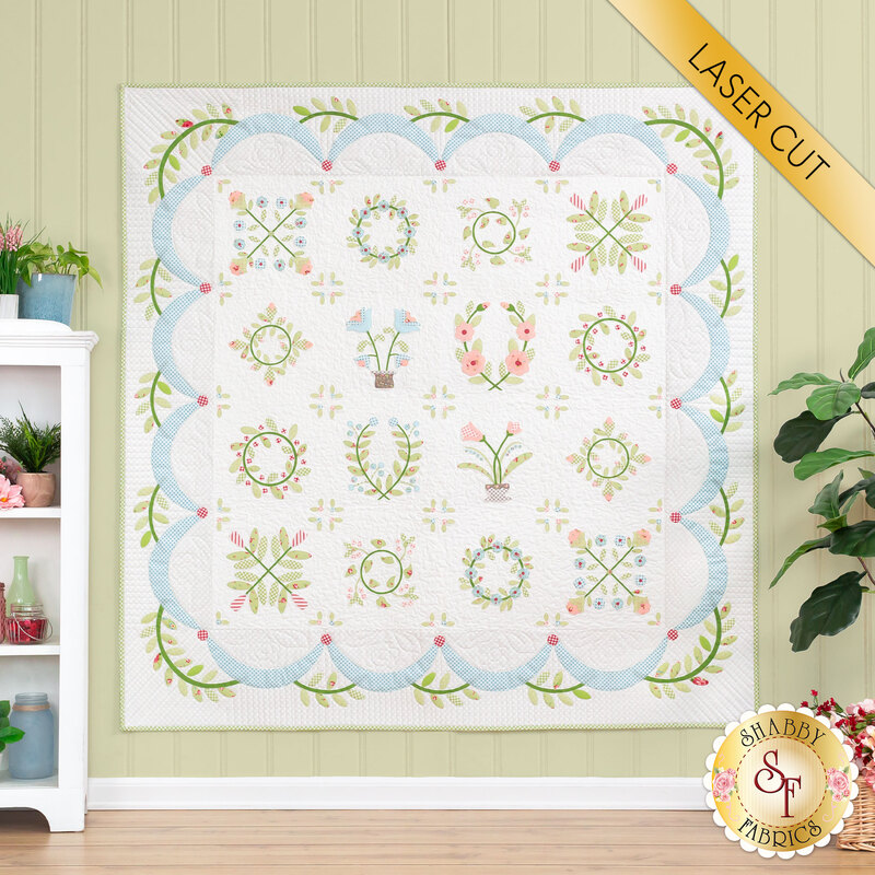 The completed Hope Blooms quilt in white, cream, blue, pink, and green, hung on a green paneled wall and staged with a house plant and white shelf that holds coordinating wares. A yellow banner reads "Laser Cut" in the upper right hand corner.