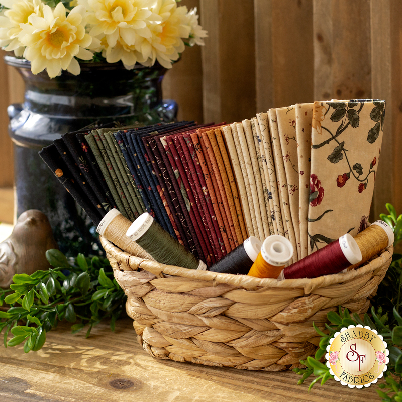 A stack of tan, golden brown, red, blue, and green fabrics set in a basket next to green sprigs, a bird figurine, and a vase of daisies