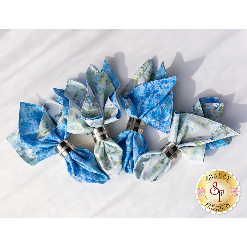 A collection of four cloth napkins in shades of blue and white, with floral patterns, arranged together. Each napkin is secured with a silver-colored clip. The background is a light, neutral color.