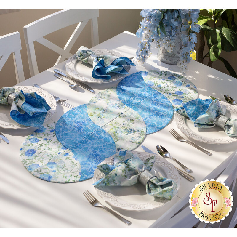 A beautifully set dining table featuring a floral table runner in shades of blue and green, with four decorative plates, each accompanied by matching napkins. The setting is bright and inviting, complemented by soft lighting and a floral centerpiece.