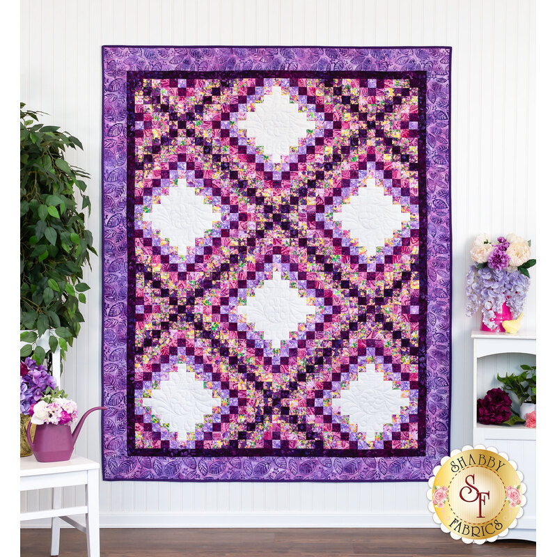 A shot of the full and completed Irish Chain quilt in bright purples, pinks, and white, hung on a white paneled wall and staged with coordinating furniture and decor.