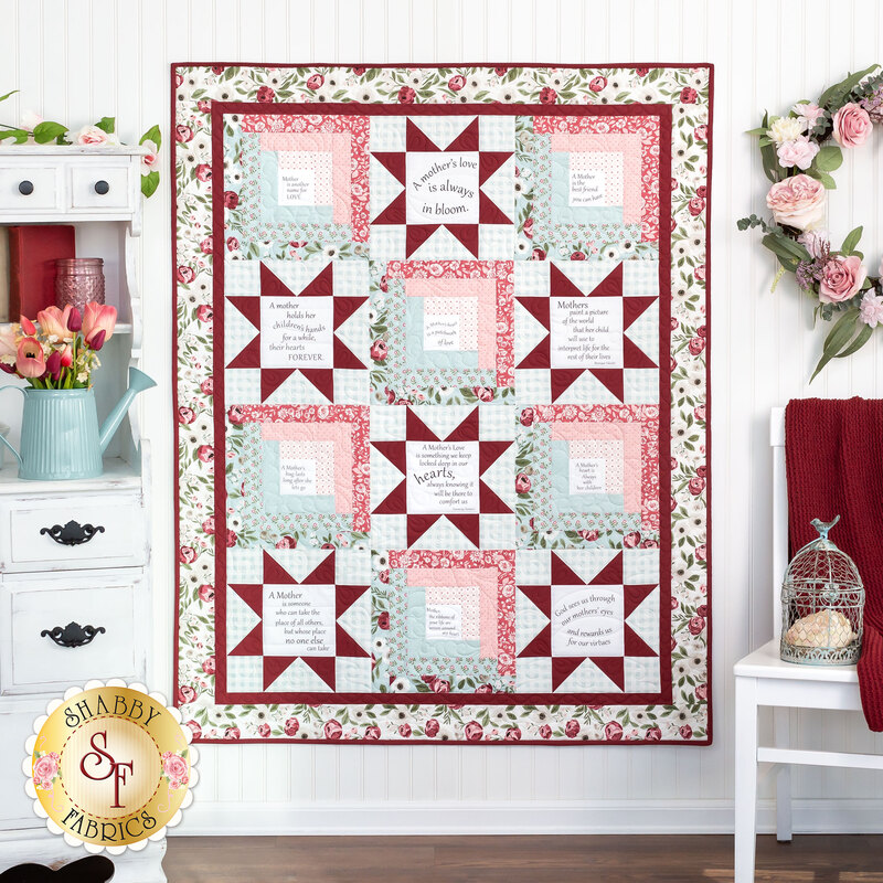 A shot of the completed Patchwork Sentiments Quilt in Lovestruck, a gentle palette of pink, powder blue, deep red, and white prints. The quilt is hung on a white paneled wall and staged with coordinating decor like furniture, flowers, and a spring wreath with pink roses.