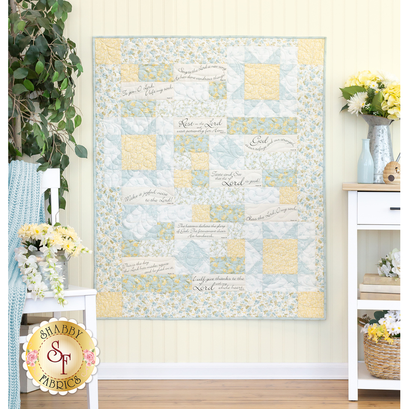 The completed Comfort of Psalms quilt in the Honeybloom collection, a palette of cream, light blue, and butter yellow in various floral prints. The quilt is hung on a white paneled wall and staged with coordinating furniture and decor.