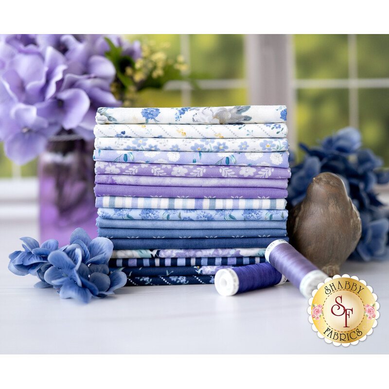 A stack of blue, purple, and white floral fabrics included in the Morning Blooms FQ Set, with flowers and a jar and thread and a bird statue.