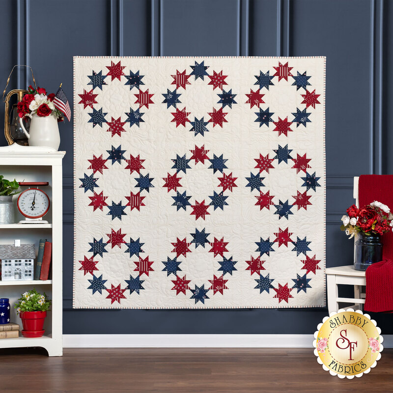 The completed Stars Above quilt, a crisp white quilt with circles of red and blue stars, hung on a cadet blue wall and staged with coordinating furniture and decor.