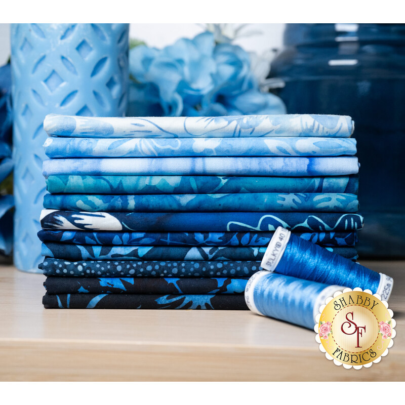 A stack of the fabrics included in the Bali Batiks-Bet on Blue collection, with flowers, a vase, a candle, and thread.