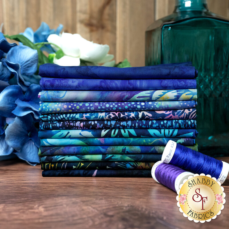 A stack of the dark navy blue and lighter blue batik fabrics included in Bali Batik Follow the Rainbow, with flowers, thread, and a glass bottle.