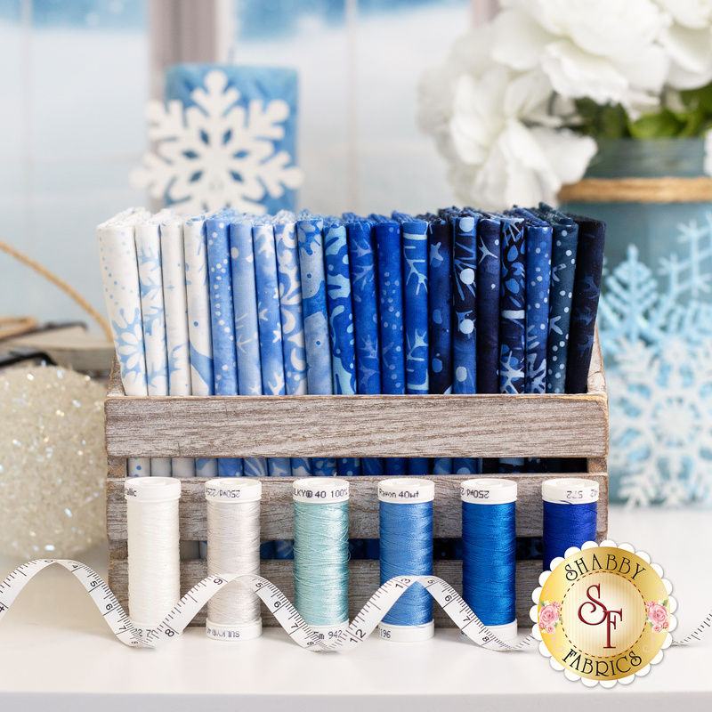 A wooden crate holds various blue fabric bolts and spools of thread, with winter-themed decor in background.