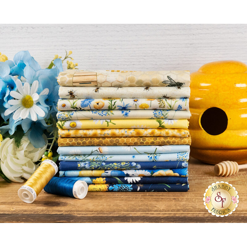 A stack of yellow, blue, and cream fabrics that are in the Bee & Bee Kind FQ set, with flowers and a ceramic beehive.