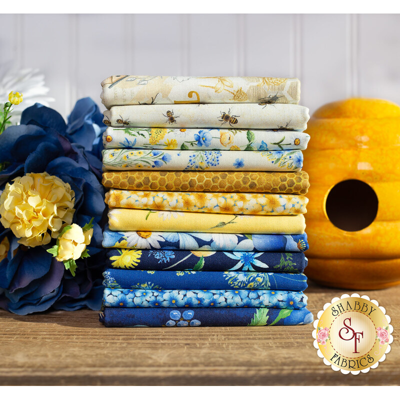 A stack of yellow, blue, and cream fabrics that are in the Bee & Bee Kind FQ set, with flowers and a ceramic beehive.