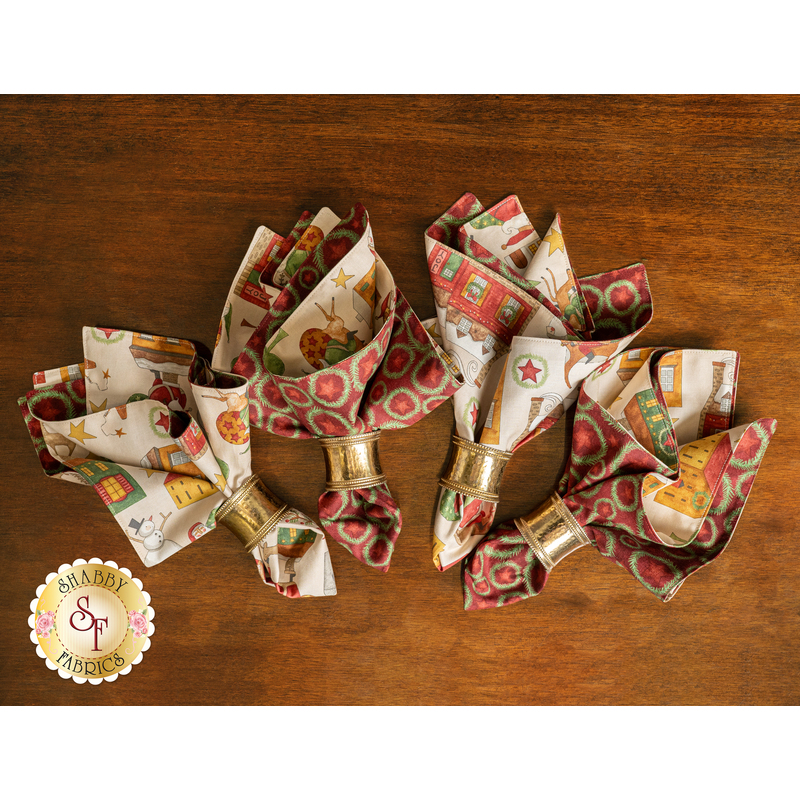 The 4 completed reversible Up On The Housetop Cloth napkins made of red and cream Christmas fabrics fanned out with gold napkin holders staged on a wooden table.