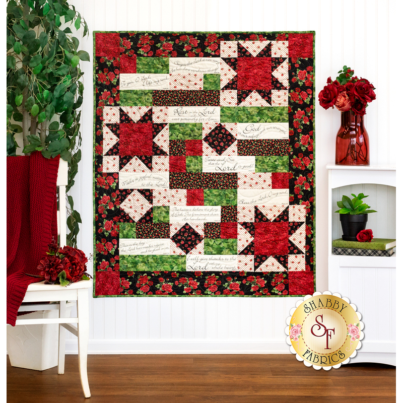 The completed Comfort of Psalms quilt in Vintage Rose, a collection of red, green, and cream floral fabrics. The quilt is hung on a white paneled wall and staged with coordinating furniture and decor, including red roses.