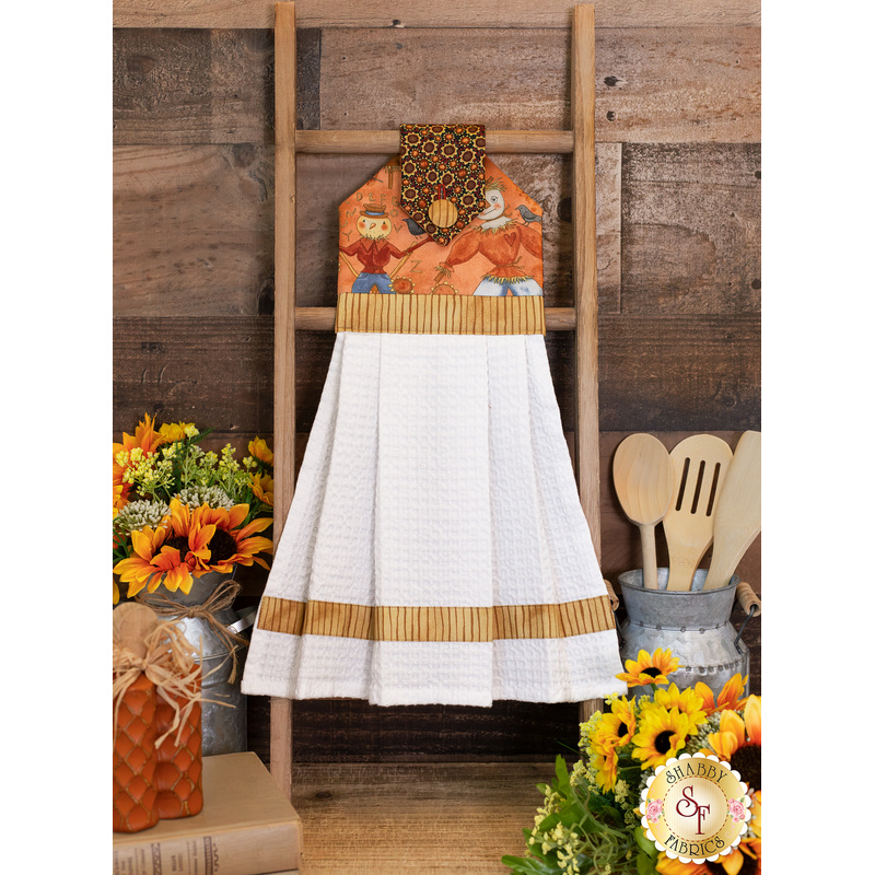 The completed hanging towel in orange, hung from a kitchen rack and staged with coordinating decor and greenery.