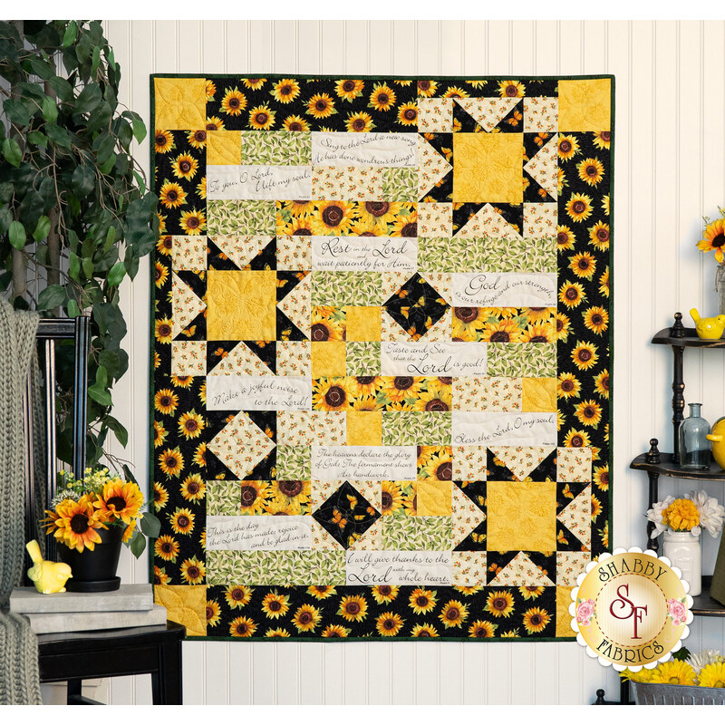 The completed Comfort of Psalms Quilt, colored in bold yellow, black, white, and green fabrics from the Sunflower Splendor collection. The quilt is displayed on an off-white paneled wall and staged with coordinating Sunflower themed decor.