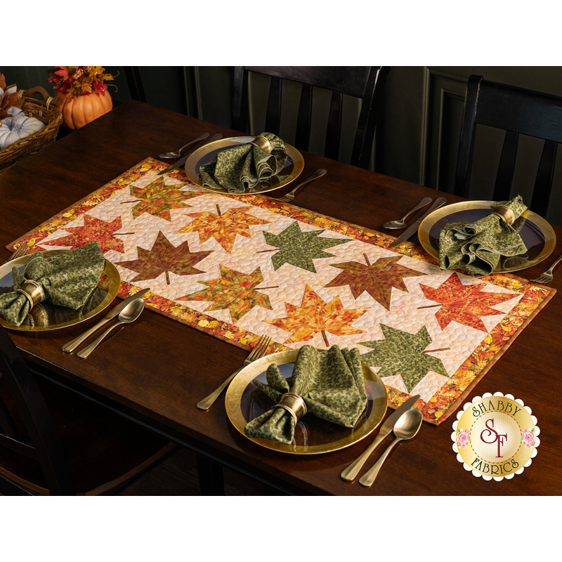 The finished table runner in Autumn Celebration displayed on a dark brown wooden table set with coordinating Autumn Celebration fabric napkins and tableware with autumnal decor peeking out of the top left corner.