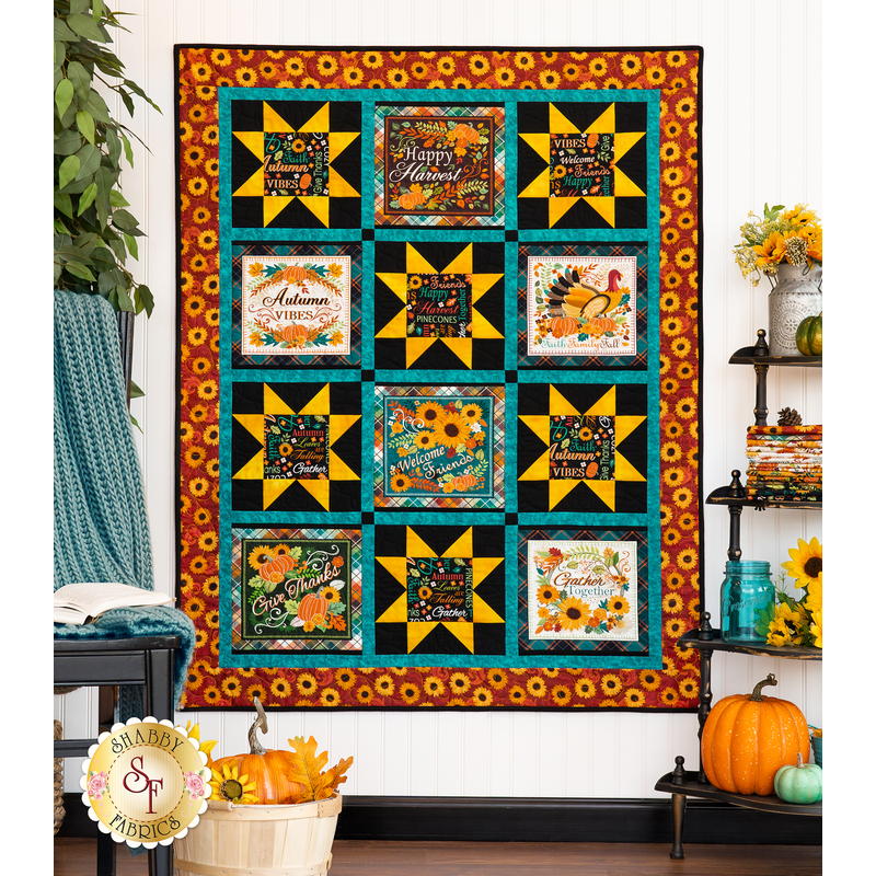 The finished Gather Together panel quilt in warm orange and teal colors, hung on a white paneled wall with a shelf on the right covered in sunflower and fall-themed decor. On the left, a leafy tree sits behind a knit blanket that is thrown over a chair.