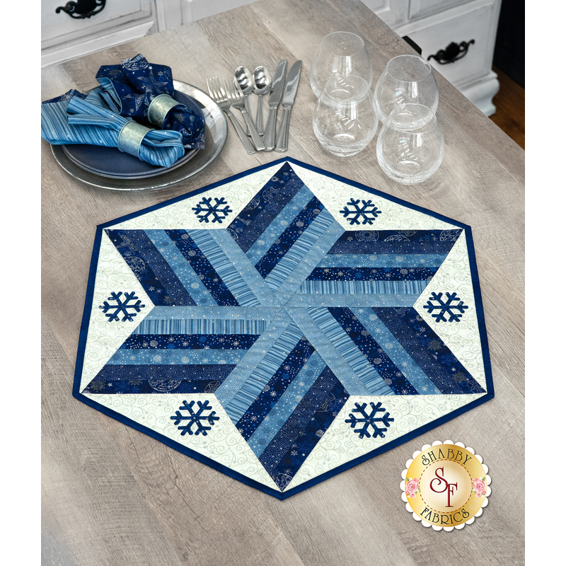 A hexagonal quilted table centerpiece project featuring winter colors and applique snowflakes, displayed on a gray table decorated in tableware.