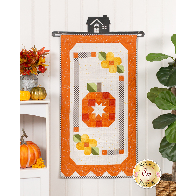 The completed October Door Banner, a lovely pumpkin with patchwork floral accents colored in bright orange, white, and yellow, hung on a white paneled wall and staged with coordinating decor.