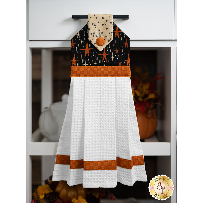 A hanging kitchen towel with an orange and black star pattern on the top and white textured fabric below, hung on a drawer handle in front of a shelf holding a teapot and decorative pumpkin.