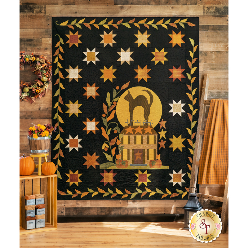 The completed Midnight Silhouette quilt, colored in bold black and warm yellow, orange, and cream fabrics, hung on a rustic wood paneled wall and staged with coordinating warm, fall decor.