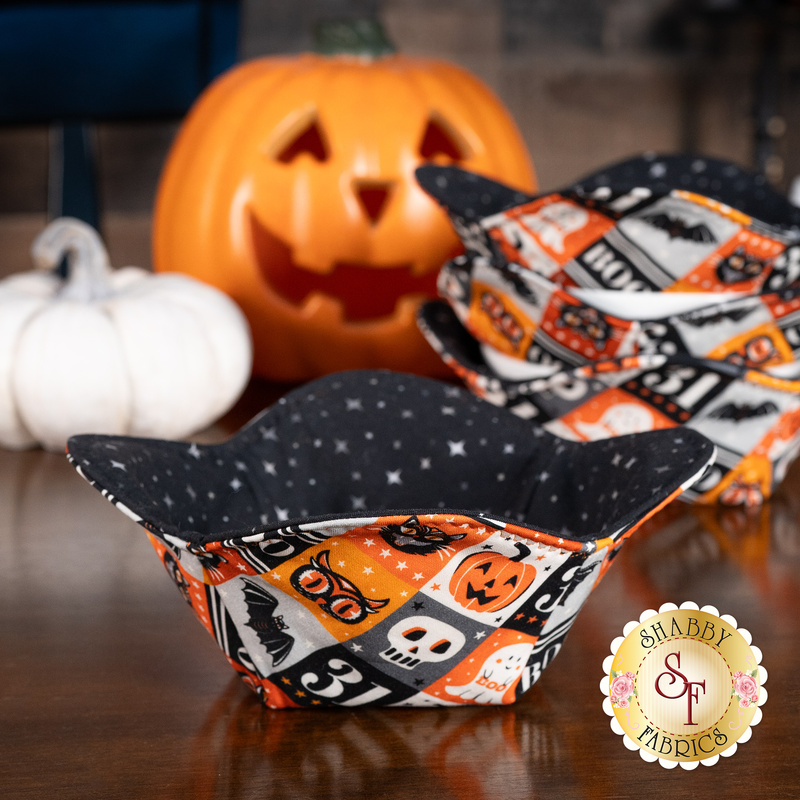 A completed bowl cozy colored in a multicolor tiled fabric on the outside and black starry interior, surrounded by coordinating Halloween table decor with three matching bowl cozies stacked in the background.