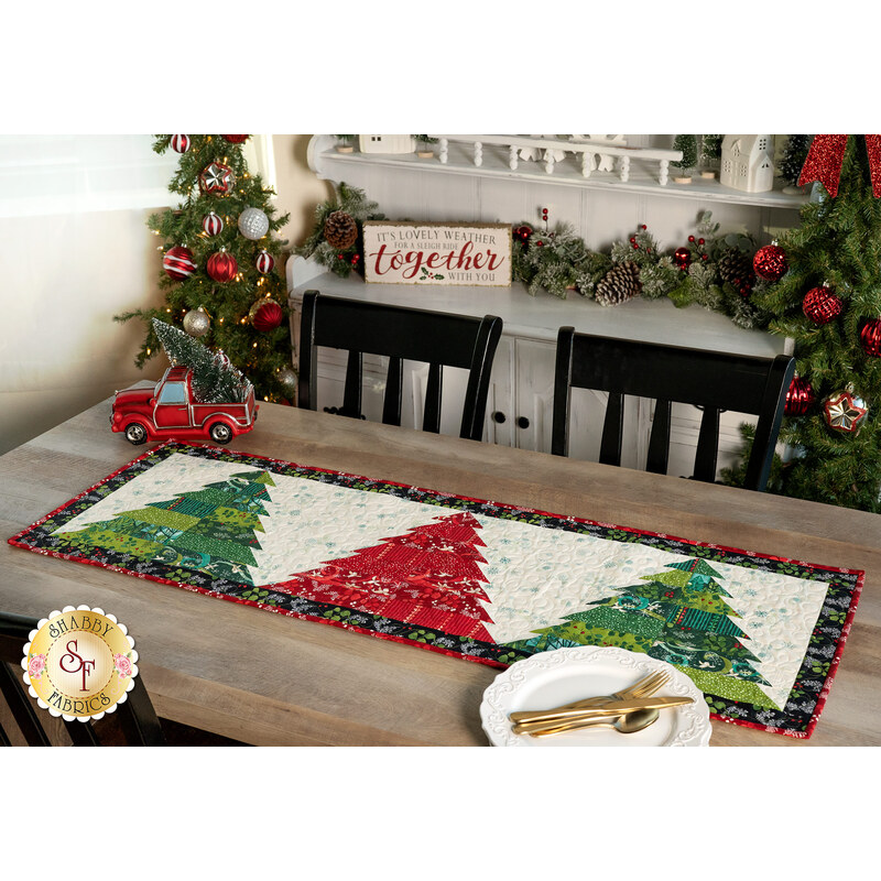 The Tree Farm Table Runner colored in red, cream, and green fabrics from the Winterly collection, staged on a rustic wood table with coordinating trees, garlands, and decor. Coordinating plates and cutlery peek into frame.