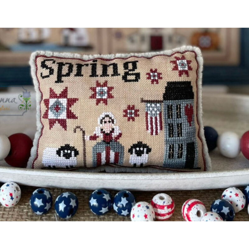 A decorative cross stitch pillow featuring the word Spring, with a farmer, sheep, and a barn in a spring theme.