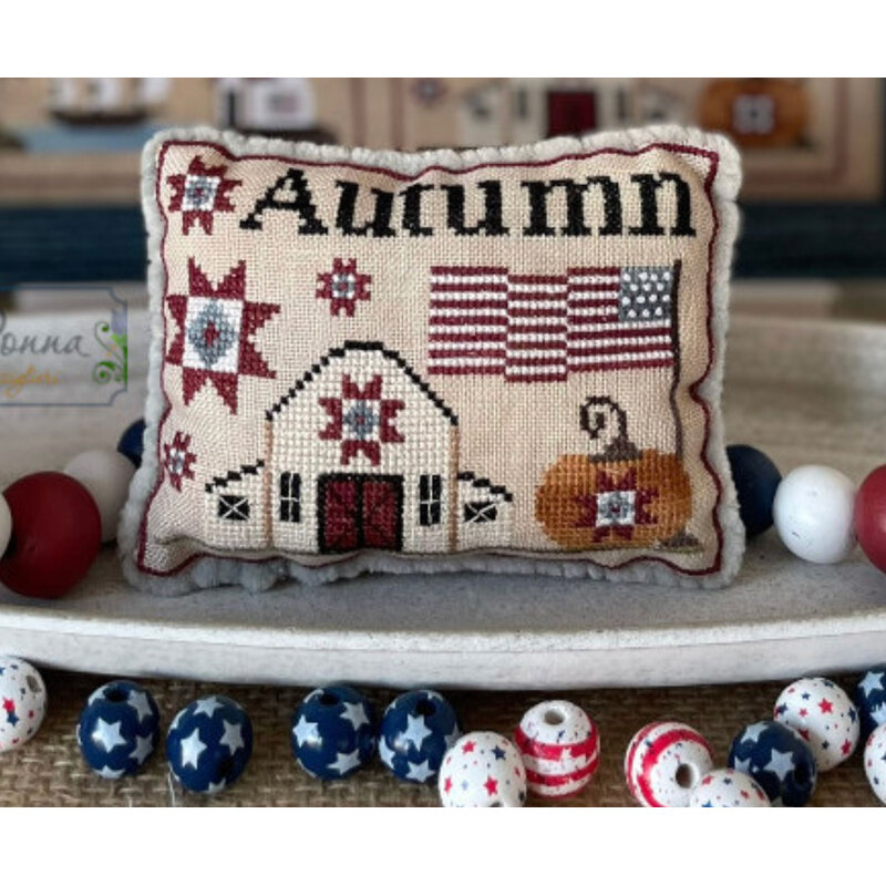 A decorative cross stitch pillow with Autumn text, featuring quilt patterns, a pumpkin, and American flags in a cozy setting.