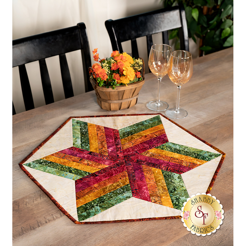 The completed 60 Degree Diamond Table Topper, colored in rich orange, red, and green fabrics from the Wine Country - Artisan Batiks collection, staged on a rustic wooden table with coordinating flowers and wine glasses.