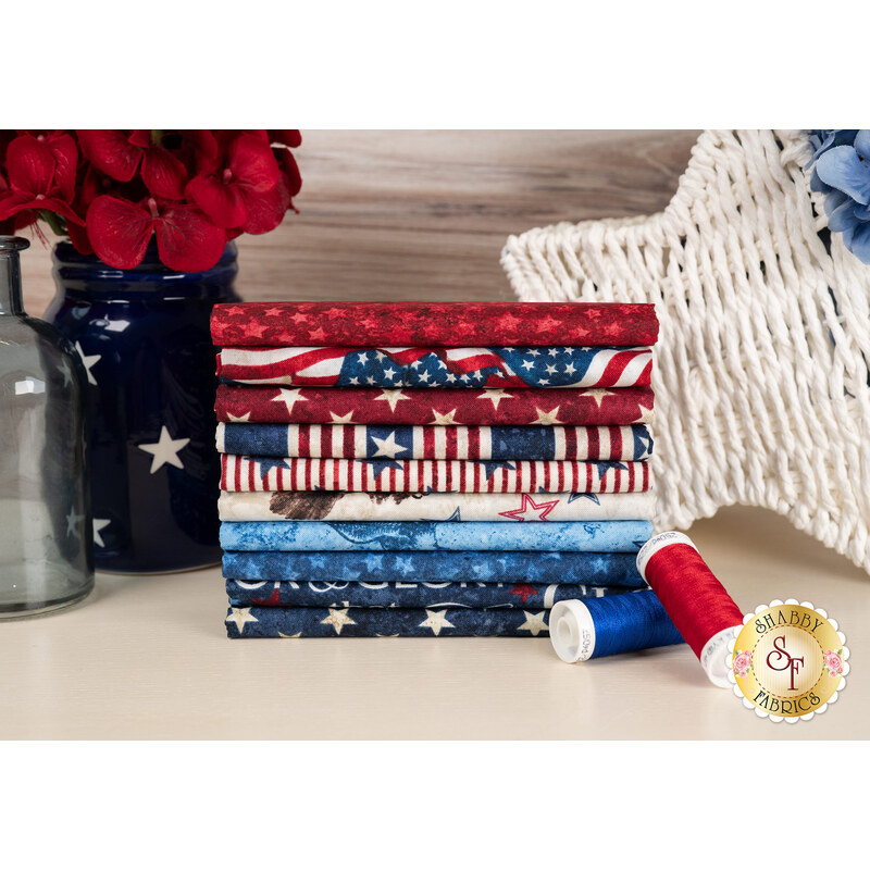 A stack of the fabrics included in the Stars & Stripes 13 FQ set, with flowers and jars and a wicker star and thread.