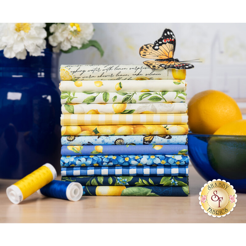 A stack of the fabric featured in the Limoncello FQ set, with lemons in a bowl and a vase and flowers and thread and a butterfly