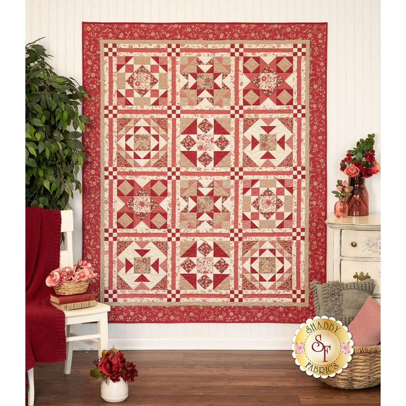 The completed Rouenneries Trois patchwork quilt, colored in rich pinks, reds, creams, and tans, hung on a white paneled wall and staged with coordinating decor.