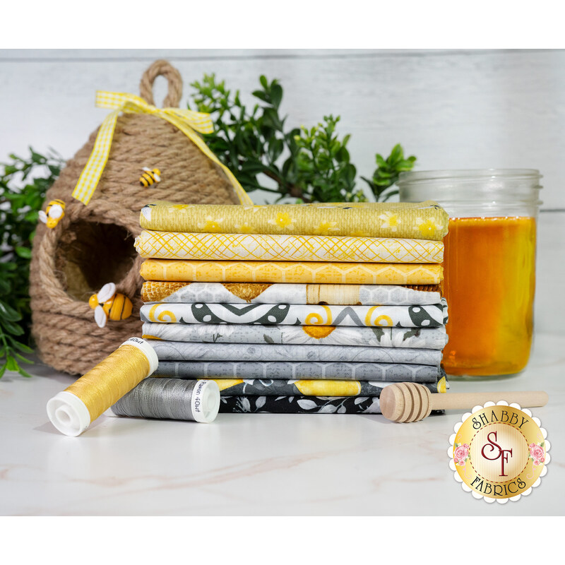 A stack of bee themed fabrics in various yellows, grays, and blacks available in the Sweet Bees 10 FQ set, with a beehive, thread, a jar of honey, and a honey dipper.