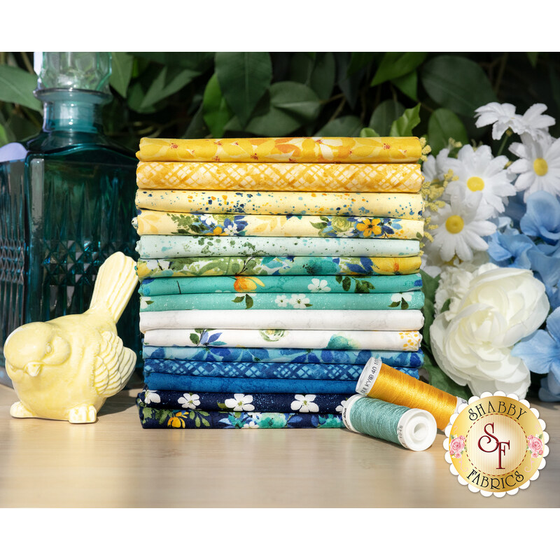 A stack of the fabrics in the Sunlit Serenade FQ set, with flowers and a glass jug and a glass bird statue and thread.