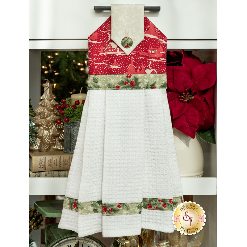 The completed towel colored in red fabric with a toile winter pattern, hanging from a kitchen drawer handle and staged with coordinating holiday decor.