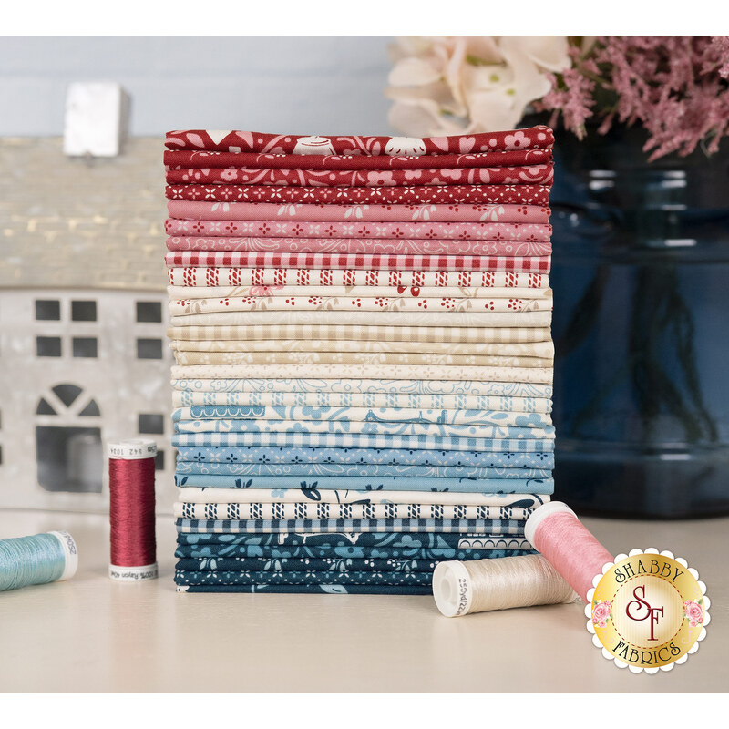 A stack of the fabrics included in the Cozy House FQ set, with a vase and flowers and thread and a house decoration.