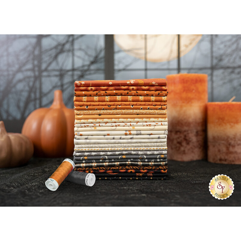 A stack of the fabrics included in the Midnight Moon collection, with a window and pumpkins and candles and thread.